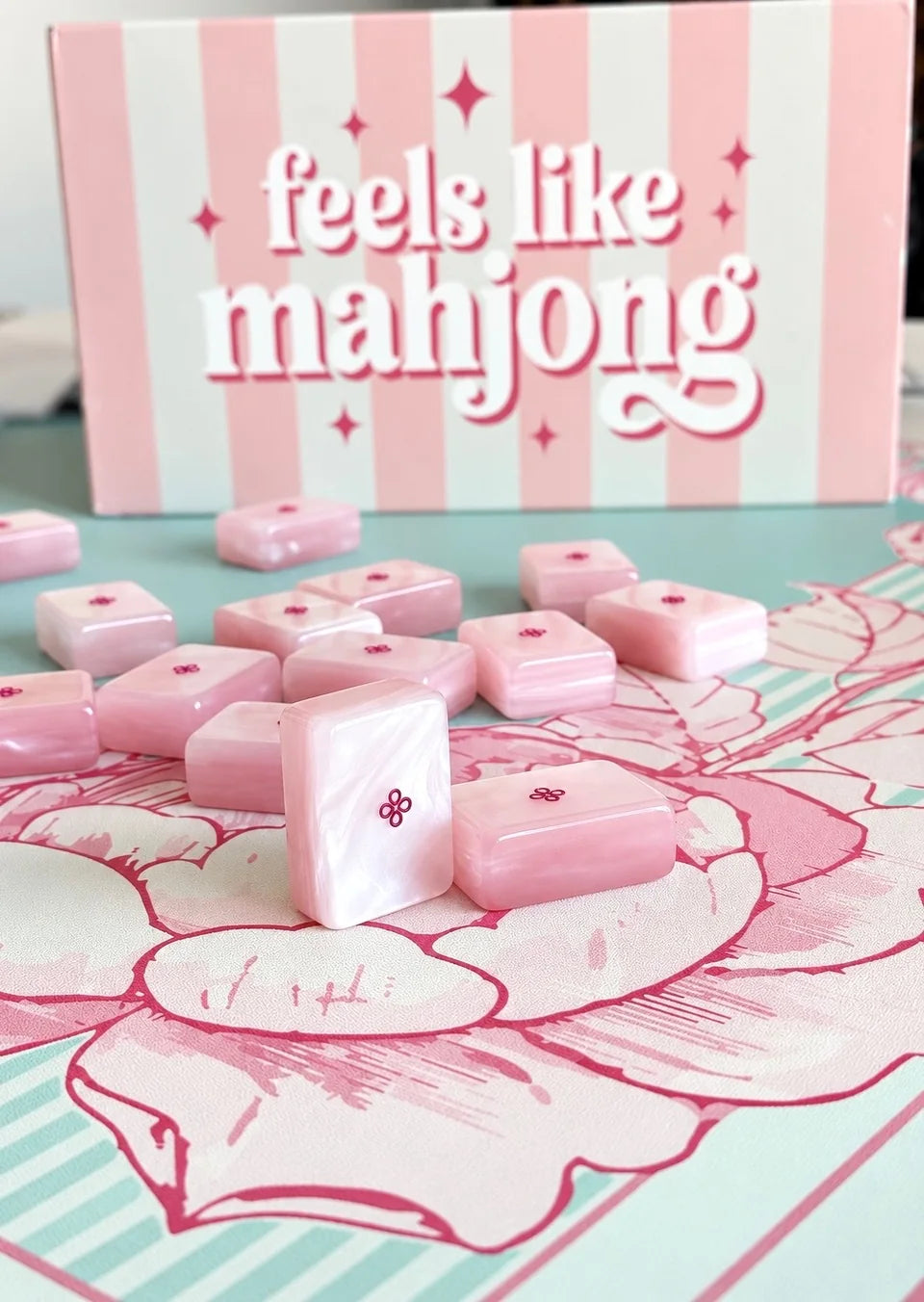 The striped feels like mahjong box standing behind pink pearl tiles, one tile upright, on a mat printed with a pink peony and mint stripes.